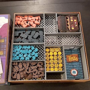 May include: A board game organiser with compartments containing game pieces in red, blue, brown, and yellow. The game box and a card with the game's title, "Tiny Towns", are also visible. The game pieces are in a variety of shapes.