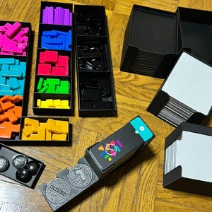 Peut inclure: Un jeu de société avec divers composants. Comprend des boîtes noires avec des formes géométriques colorées, des cartes et des pièces de jeu. Les formes sont roses, bleues, oranges, jaunes, vertes et violettes. Les pièces sont noires et argentées.
