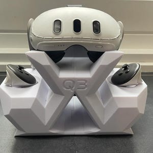 May include: A white virtual reality headset rests on a white geometric stand labeled with the text Q3. Two black and white handheld controllers are docked in the side slots of the stand, which features a faceted, angular design.