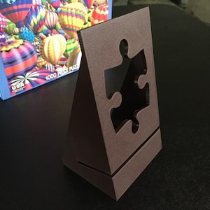 May include: A brown cardboard puzzle holder with a cut-out in the shape of a puzzle piece. The holder is angled and has a small base.