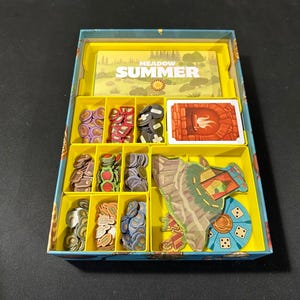 May include: A yellow and blue game organizer with compartments for storing game pieces. The organizer has a label that says "Meadow Summer".