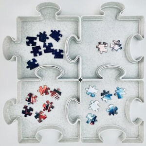 May include: A light gray puzzle organizer shaped like a large puzzle piece, divided into four sections. Each section holds a small collection of puzzle pieces with various colorful designs, including space and flag themes.