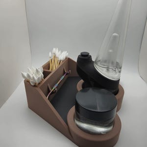 May include: A brown organizer stand featuring a black base, a circular cutout holding a black and clear glass device, a circular cutout holding a small black jar, and two rectangular compartments containing white cotton swabs and a small metal tool.