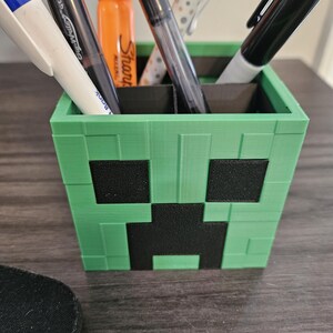 Puede incluir: Porta-bol&iacute;grafos de pl&aacute;stico verde y negro con forma de cara de Creeper de Minecraft. El porta-bol&iacute;grafos tiene cinco ranuras para bol&iacute;grafos y l&aacute;pices.