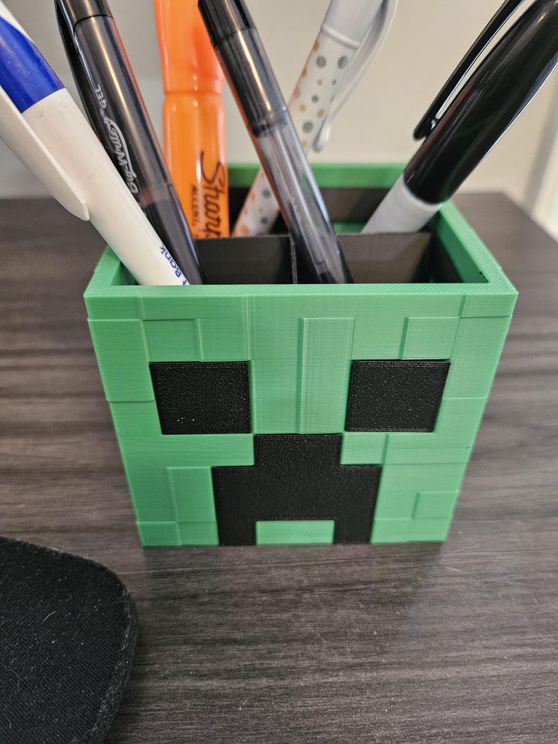 Puede incluir: Porta-bol&iacute;grafos de pl&aacute;stico verde y negro con forma de cara de Creeper de Minecraft. El porta-bol&iacute;grafos tiene cinco ranuras para bol&iacute;grafos y l&aacute;pices.