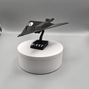 May include: A black F-117 Nighthawk model airplane, mounted on a black stand with the text "F-117". The model has a distinctive angular design and is displayed on a white rotating platform. The model is a miniature replica of the stealth fighter jet.