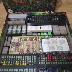 Puede incluir: Un tablero de juego negro y verde con varios compartimentos para guardar las piezas y las cartas del juego. El tablero tiene una etiqueta que dice "rebel".