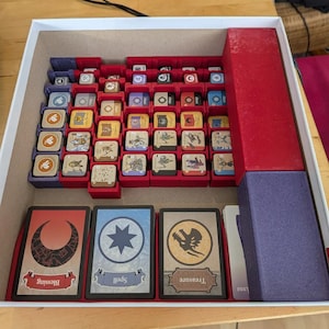 May include: A board game box filled with game components. The box contains red trays holding various game pieces, including small tokens and cards labeled "Blessing," "Spell," and "Treasure." The box is open, revealing the organized contents.