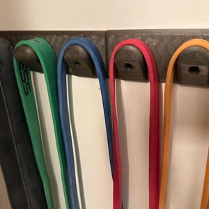 May include: A wall-mounted rack with five resistance bands hanging from it. The bands are black, green, blue, red, and orange.