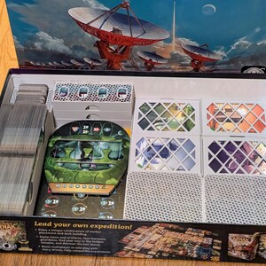 May include: Open board game box with various components. Includes stacks of cards, game tiles, and a central game piece with a green and brown design. The box art features a space-themed illustration with satellite dishes and a planet.