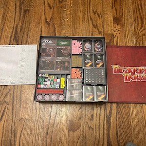 May include: An open board game box with various components, including cards, dice, and tokens. The game is titled "Dragons Down" on the red game board cover. The game pieces are organized in a black tray.