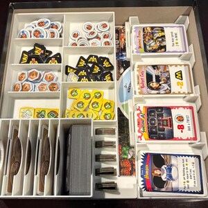 May include: A white plastic game organizer filled with various game components. The organizer has multiple compartments holding tokens, cards, and other game pieces. The cards have text like "Hunger" and "Budget Cuts". The game pieces are in various colors.