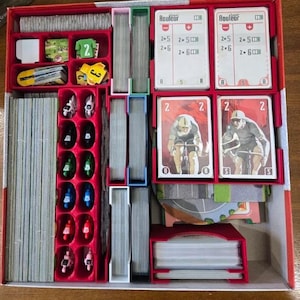May include: A red board game organizer filled with game components. It contains cards, player pieces, and other game elements. The cards feature cyclists and the word "Rouleur". The organizer is designed to keep the game pieces organized.