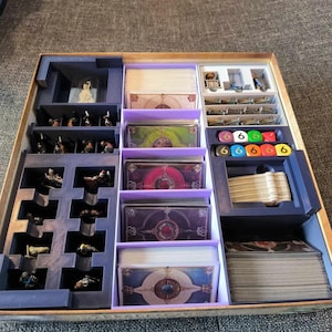 May include: A wooden game organizer with multiple compartments. It contains card decks, miniature figures, dice, and other game components. The organizer is primarily blue and purple, with a light brown wooden frame.