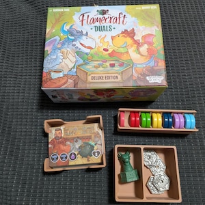 May include: A board game box titled "Flamecraft: Duels Deluxe Edition" with colorful dragon illustrations. Includes game pieces in brown trays, such as colored tokens and a green dragon figurine. The game box is on a dark gray surface.