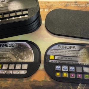 May include: Black game pieces with white text and graphics. The pieces are labeled "Triton", "Ganymede", and "Europa". The game pieces have slots for tracking game progress. The game pieces are for a board game.
