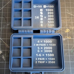 Op de afbeelding: Open blauwe plastic dobbelspelbox met compartimenten en tekst. De box is open en onthult twee helften met meerdere compartimenten voor het opbergen van dobbelstenen. Tekst bevat dobbelsteencombinaties en puntwaarden.