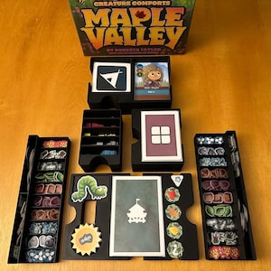 May include: A board game titled "Maple Valley" with various game components, including cards, tokens, and organizers. The game pieces are arranged on a wooden surface, showcasing the game's design and components.