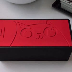 May include: A rectangular black box with a vibrant red lid featuring an embossed line drawing of a cat face. The cat design includes large circular eyes, a small nose, and whiskers. The box has a matte finish and a clean, geometric structure.
