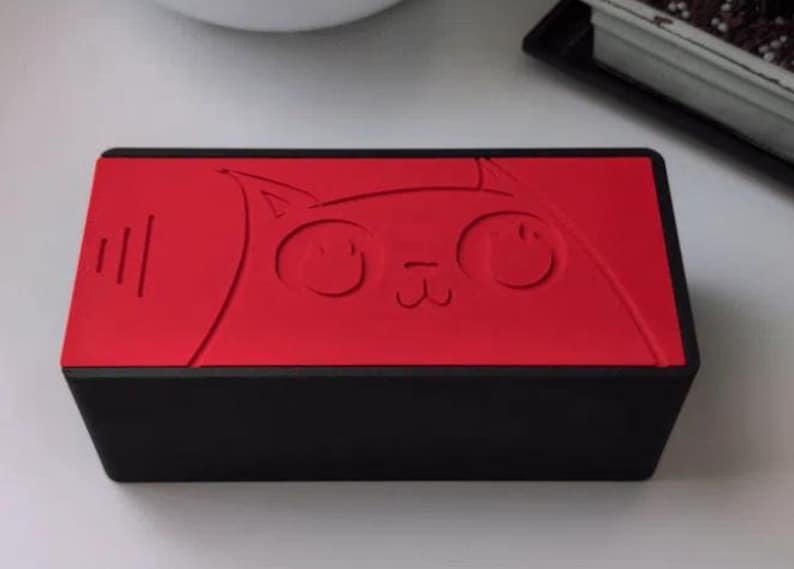 May include: A rectangular black box with a vibrant red lid featuring an embossed line drawing of a cat face. The cat design includes large circular eyes, a small nose, and whiskers. The box has a matte finish and a clean, geometric structure.