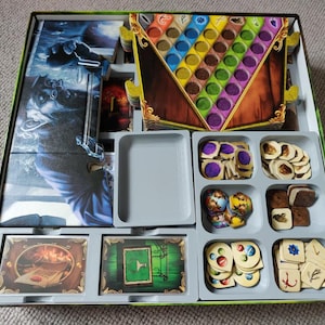 Puede incluir: Un organizador de juegos de mesa gris y blanco con múltiples compartimentos para guardar las piezas del juego. El organizador tiene un compartimento para un tablero de juego grande y colorido con un marco de madera. El organizador también tiene compartimentos para piezas de juego más pequeñas, como fichas, dados y cartas.