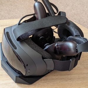 May include: A black virtual reality headset with a glossy front visor, adjustable fabric head straps, and two handheld controllers resting on a black charging dock. The headset features small buttons on the side and a textured face interface.