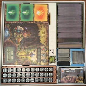 May include: A board game box with various components. The box contains cards, dice, and game boards with illustrations. The game boards feature the text "HILDA" and "Fable Fury". The dice are gray with white dots. The box is organized with dividers.
