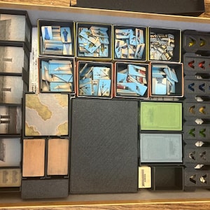 May include: A board game organizer with various compartments filled with game components. The components include cards with images of ships and landscapes, along with other game pieces. The organizer is made of cardboard and plastic.