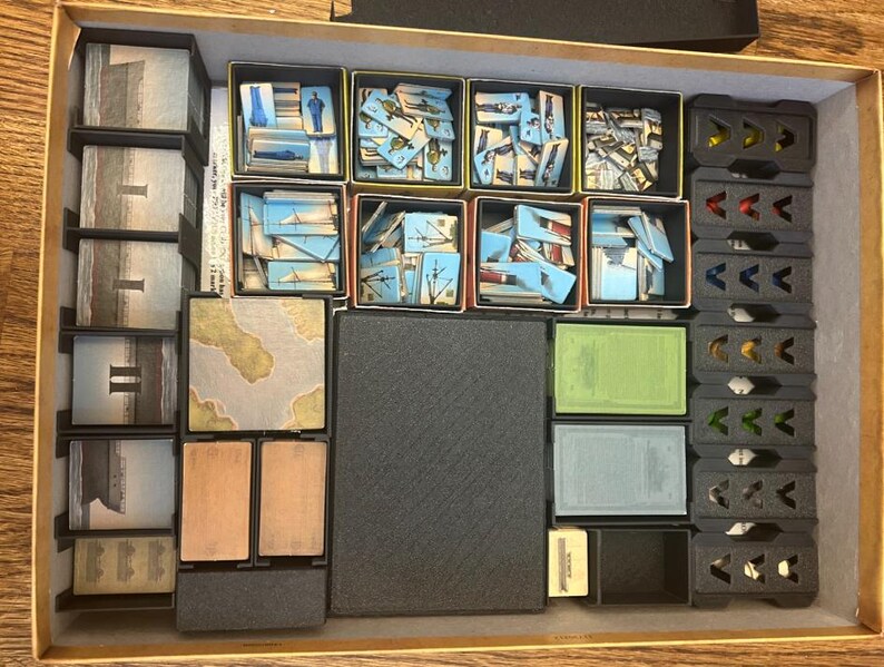May include: A board game organizer with various compartments filled with game components. The components include cards with images of ships and landscapes, along with other game pieces. The organizer is made of cardboard and plastic.