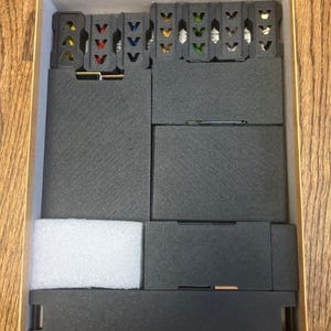 May include: A cardboard box with a wooden grain pattern, containing black foam inserts and several small, dark gray plastic components with colored accents. The box is open, revealing the organized contents.