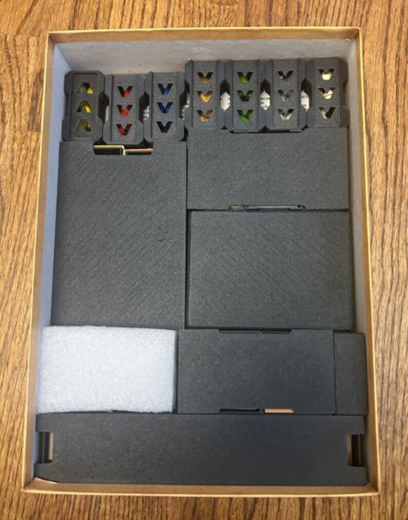 May include: A cardboard box with a wooden grain pattern, containing black foam inserts and several small, dark gray plastic components with colored accents. The box is open, revealing the organized contents.