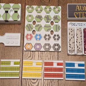 Puede incluir: Un juego de mesa con varios componentes, incluyendo fichas hexagonales, fichas, dados y un panel de control. Las piezas del juego son en tonos de verde, rojo, azul, amarillo y gris. La caja del juego tiene el texto "Age of Steam".