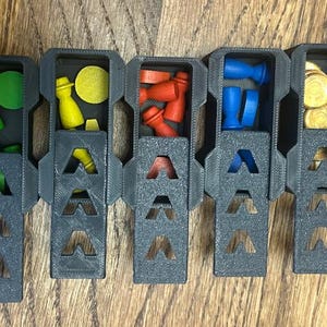 May include: A segmented, dark gray game piece organizer with various colored game pieces. The organizer contains wooden pieces in white, green, yellow, red, blue, and gold. The organizer is on a wooden surface.