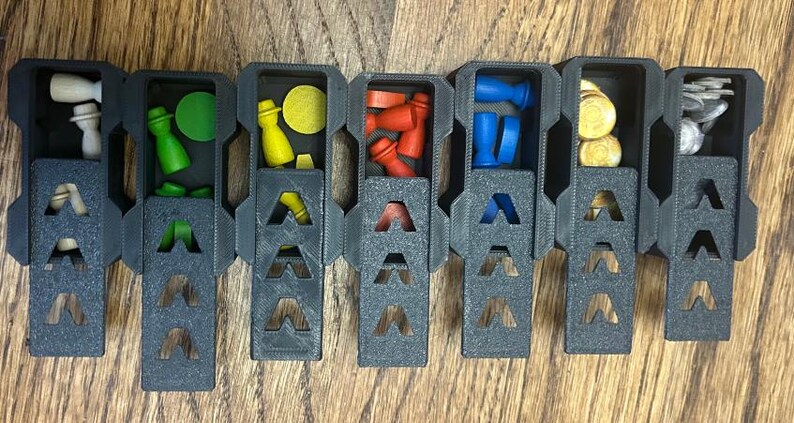 May include: A segmented, dark gray game piece organizer with various colored game pieces. The organizer contains wooden pieces in white, green, yellow, red, blue, and gold. The organizer is on a wooden surface.