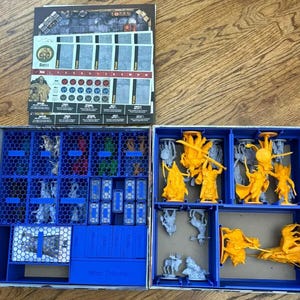 May include: A board game box organizer featuring blue plastic inserts with a honeycomb pattern. The left side contains small compartments for tokens and cards, while the right side holds yellow and grey miniature figures. A game board sheet is included.