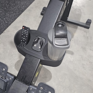 May include: A black rowing machine with a built-in accessory tray. The tray holds a remote control, a water bottle, and a small device. The machine has footrests and a handle. The background is a gray floor and a black mat.