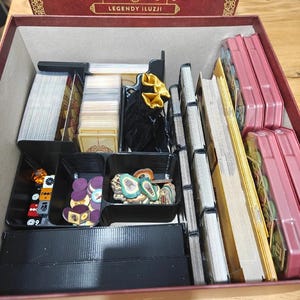 May include: An open box game organizer for the board game "Trickerion: Legends of Illusion". The organizer is made of black plastic and has compartments for cards, tokens, and dice. The game box is red with gold lettering and the title "Trickerion: Legends of Illusion".