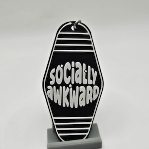 Puede incluir: Llavero de motel negro y blanco con el texto "socially awkward" en letras blancas.