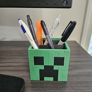 Puede incluir: Un portal&aacute;pices verde en forma de Creeper de Minecraft con ojos y boca negros. El portal&aacute;pices est&aacute; lleno de bol&iacute;grafos y marcadores, incluyendo un bol&iacute;grafo blanco con lunares negros, un bol&iacute;grafo negro, un bol&iacute;grafo azul, un marcador negro y un resaltador naranja.