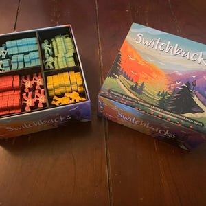 May include: A box game called Switchbacks with a colorful mountain scene on the lid. The box is open and shows the game pieces, including small wooden figures and colored tiles.