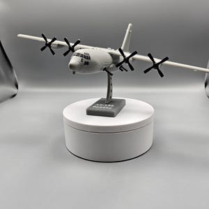 May include: A gray model airplane with four propellers on each wing and a detailed cockpit. The model is mounted on a stand that reads "AC-130 Spooky". The airplane is displayed on a white, circular platform.