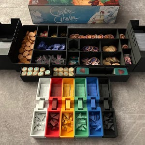 May include: A black organizer with multiple compartments for storing game pieces. The organizer is filled with various colored plastic pieces, including red, blue, green, yellow, and white. The organizer is labeled "Creature Caravan".