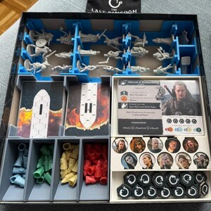 May include: A board game box organizer containing gray plastic miniature figures in blue dividers, stacks of rectangular game cards, and four compartments holding blue, green, yellow, and red plastic tokens. A character card for Uhtred of Bebbanburg is visible alongside circular character portraits and black hexagonal tokens.
