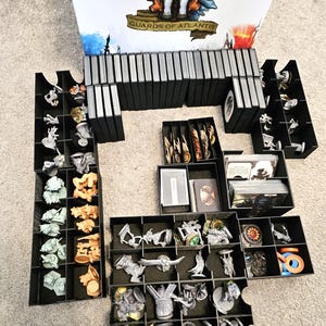 May include: A black plastic organizer with multiple compartments filled with miniature figures and game pieces. The organizer is labeled "Guards of Atlantis".