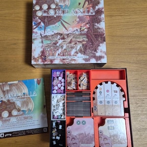 May include: A board game box set with the title "COMPANIA" in a stylized font. The box art features a fantasy landscape with airships. Inside, an orange organizer holds game components, including cards and tokens. An instruction manual is also visible.