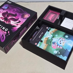 May include: A Casting Shadows board game set featuring a black box with purple and pink character graphics, and an open game tray containing a deck of cards, a rulebook, small colored character tokens, and a game board with an axolotl illustration.