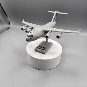 Puede incluir: Maqueta gris de un avión C-17 Globemaster III, expuesta sobre un soporte circular blanco. El modelo tiene cuatro motores y alas detalladas. El soporte tiene una base gris con el texto "C-17 Globemaster III".