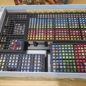 May include: A blue and white game box with a black foam insert for organising game pieces. The insert has multiple compartments for storing cards, tiles, and other game components.