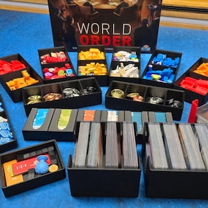 May include: A board game titled "World Order" with various game pieces and card decks. The game pieces are organized in black trays, with red, yellow, orange, and blue components. The game also includes gold and silver coins, and a red card holder.