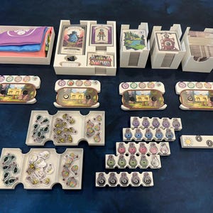 May include: A collection of board game components, including white storage boxes, cards, tokens, and game boards. The components are arranged on a dark blue surface. The game pieces feature various colors and designs, suggesting a fantasy or adventure theme.